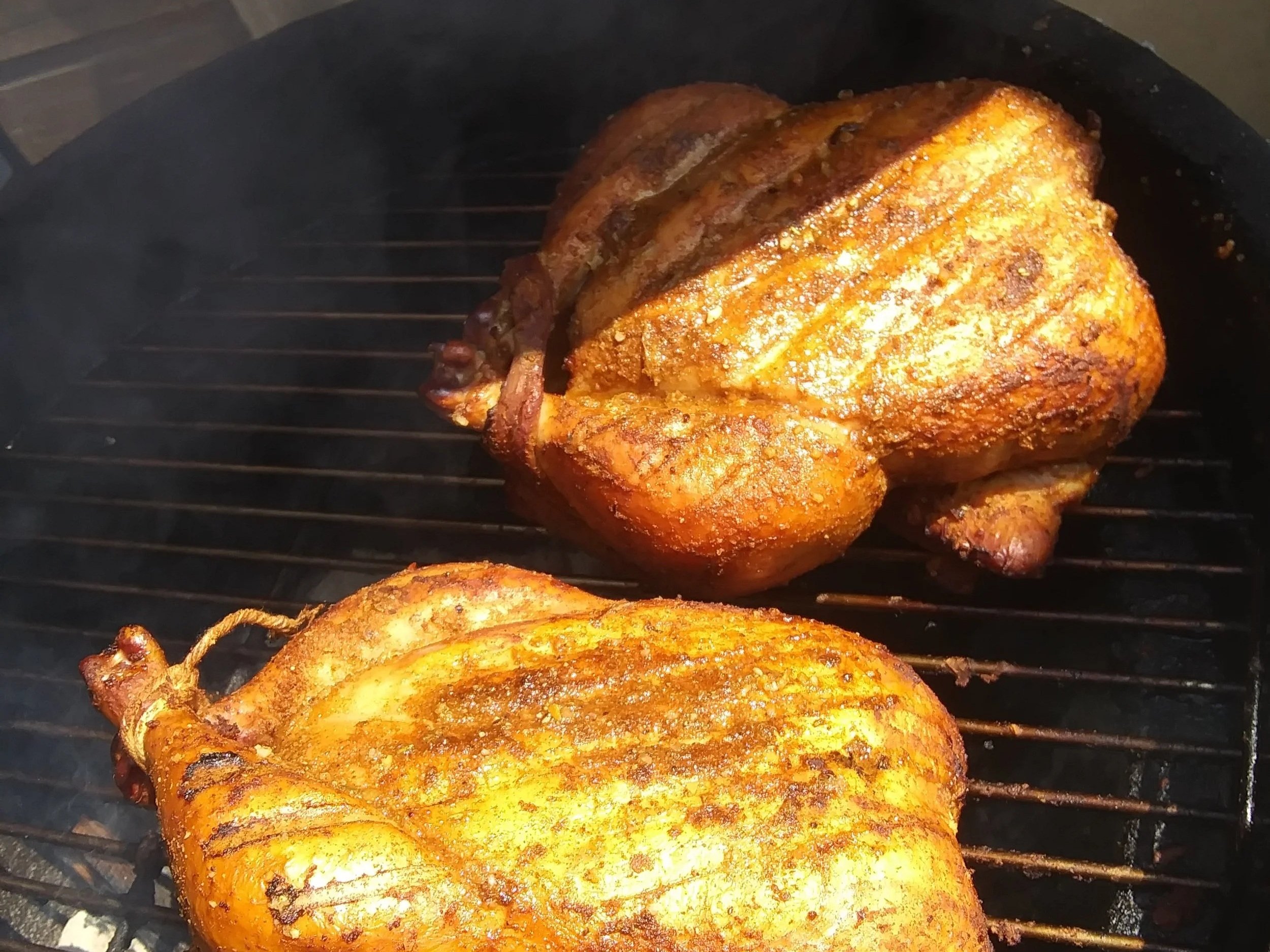 Two perfectly grilled whole chickens with a crispy, golden-brown skin and visible grill marks, cooking on a barbecue grill with smoke rising in the background. Ideal for outdoor cooking enthusiasts and barbecue lovers.