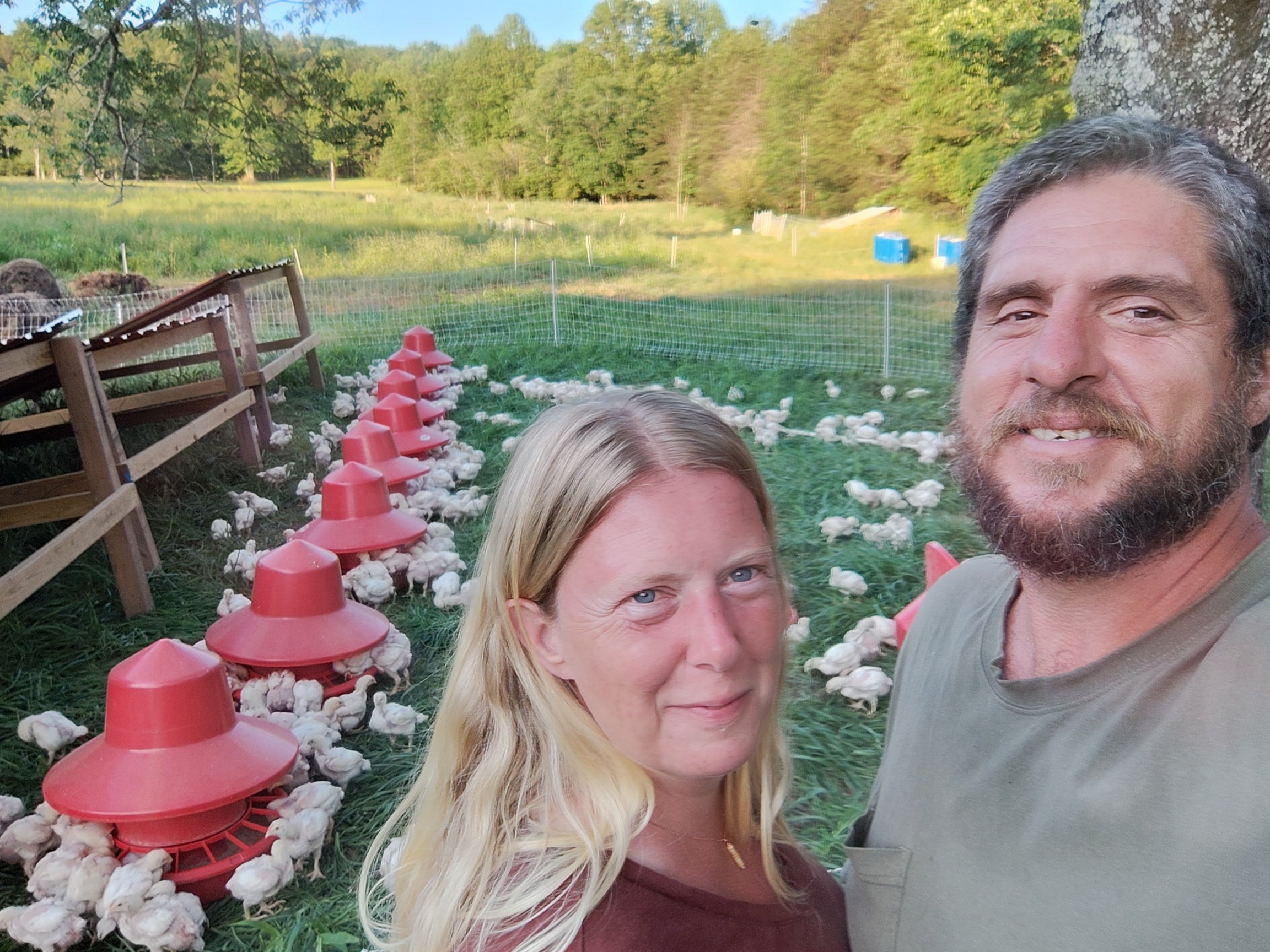 IMG_20240521_193329526_HDR Couple posing in a rural farm setting with chickens feeding near red feeders, surrounded by lush greenery and trees in the background, highlighting sustainable farming practices.