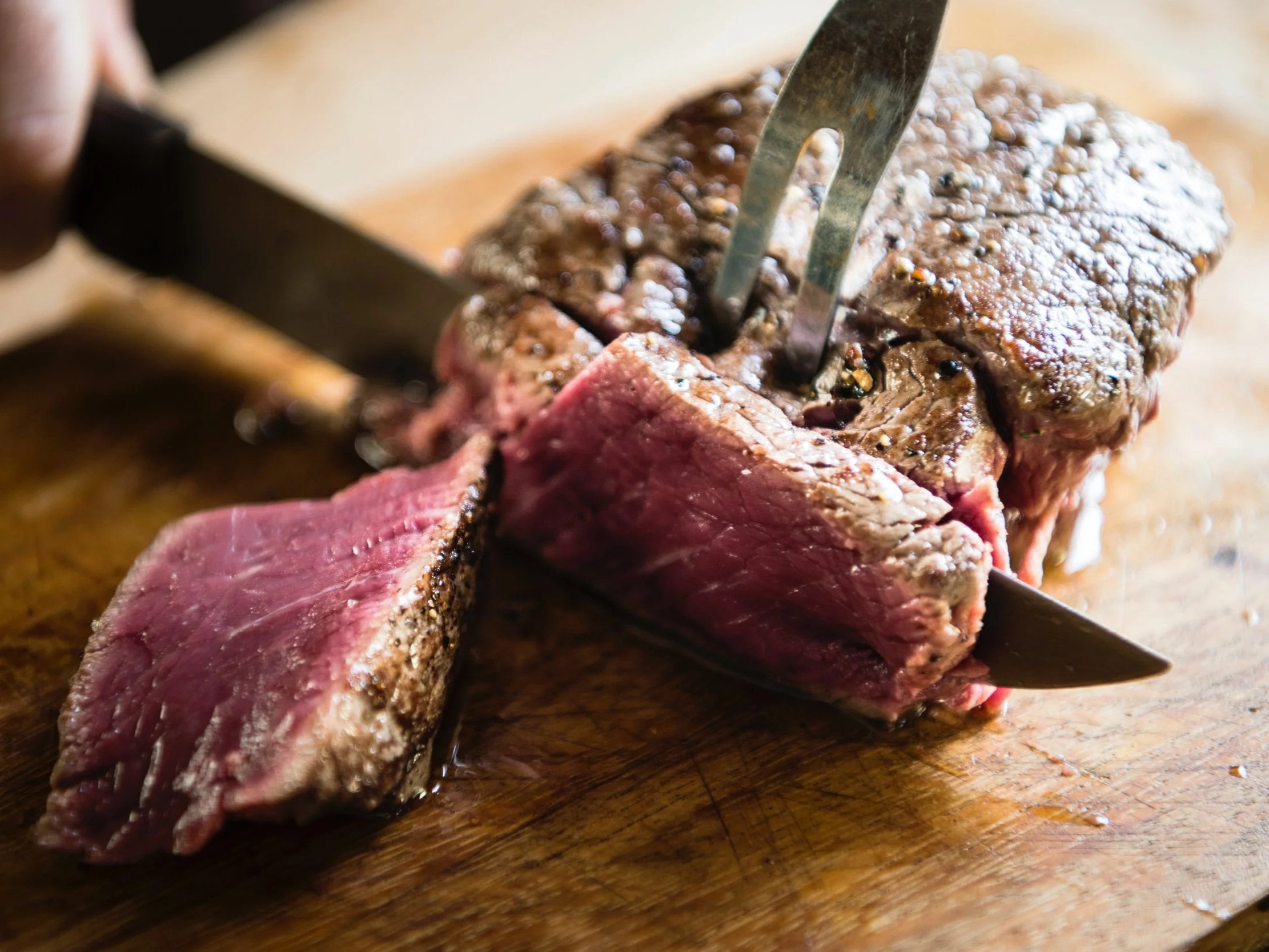 Juicy steak being sliced on a wooden cutting board, showcasing its tender pink interior and charred exterior, with a knife and fork in action.