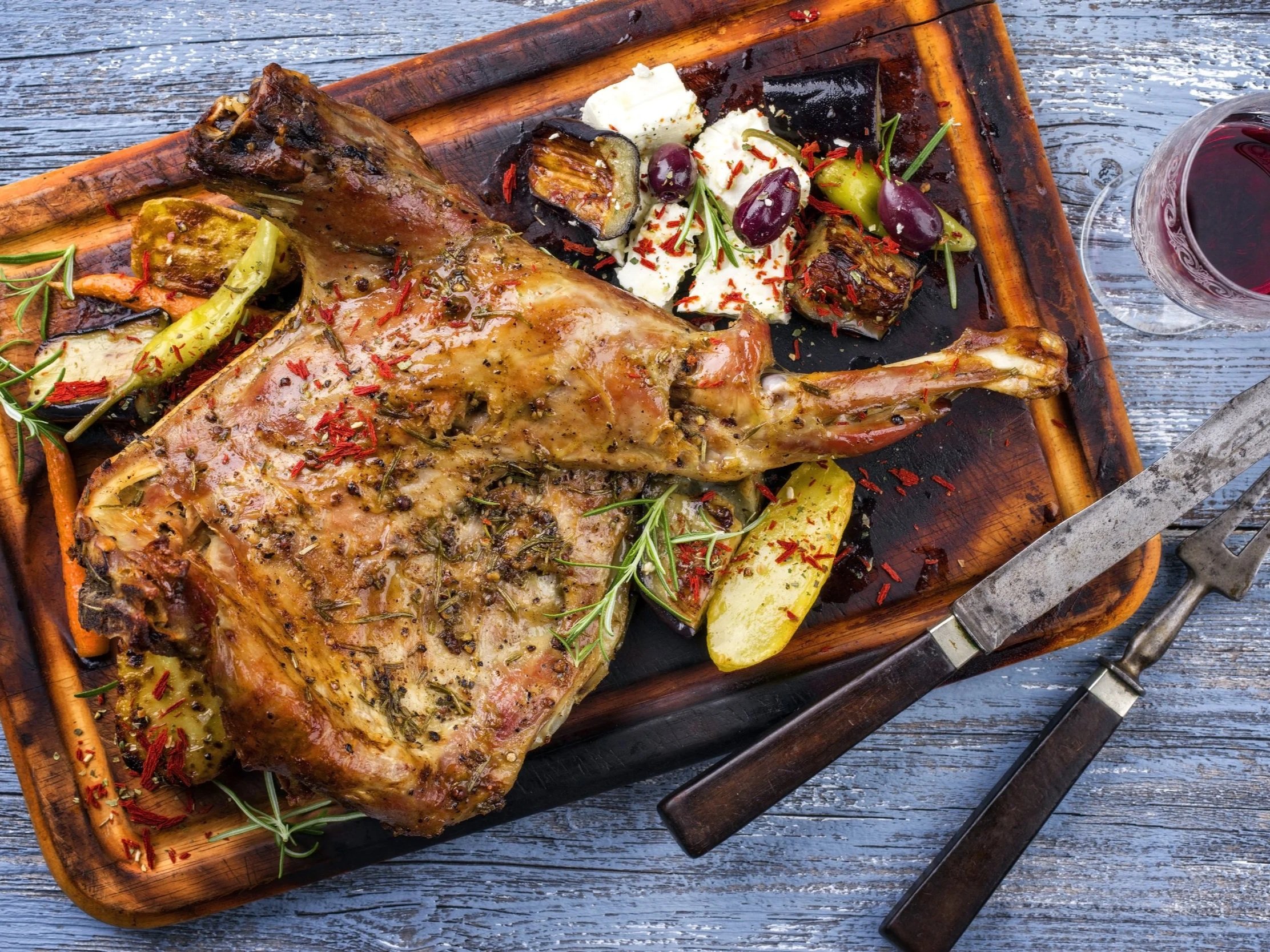 Roasted leg of lamb served on a wooden platter with grilled vegetables, garnished with herbs and spices, accompanied by a glass of red wine. Perfect for a festive meal or special occasion.